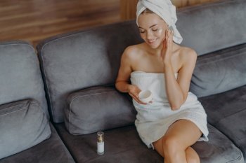Happy woman in towel applying facial cream, enjoying home spa day