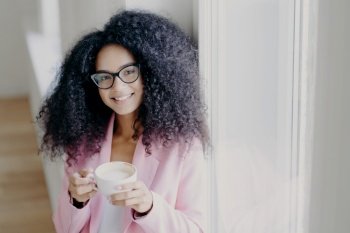 Smiling woman with curly hair enjoying a cup of coffee in a pink blazer.