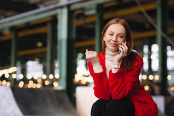 Smiling woman in red cardigan talks on phone, blurred lights in background
