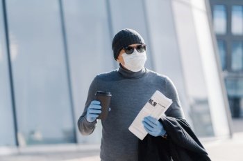 Man in winter gear with coffee and newspaper, showcasing pandemic life with gloves and mask, against a modern architectural backdrop