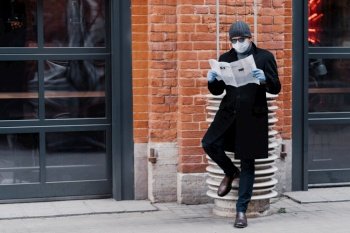 Man in a stylish winter outfit leaning against a column reading a newspaper, masked and gloved for safety, urban setting