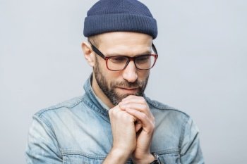 Thoughtful man in denim with hand on chin, deep in contemplation, wearing a beanie and glasses, against a neutral background