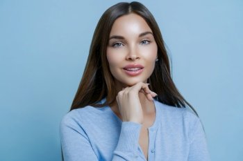 Thoughtful brunette woman in blue cardigan, hand on chin, with a subtle smile, against a pale blue backdrop