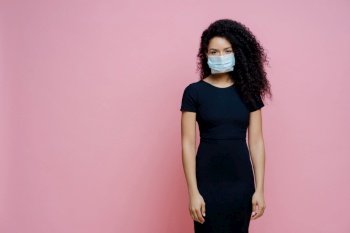 Solemn woman in black dress and face mask, pink background.