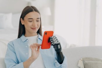Smiling young woman with prosthetic arm using smartphone, experiencing accessibility technology at home