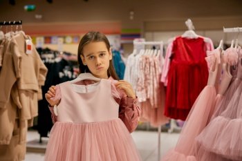 Portrait of charming little girl choosing elegant dress at shopping mall. Small fashionista shopaholic standing over clothing displayed on rack. Portrait of charming little girl choosing elegant dress at shopping mall