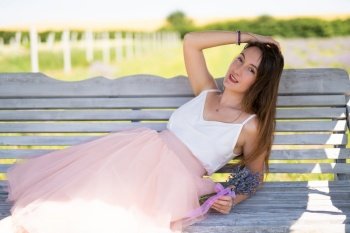 a beautiful girl in a dress with a bouquet of lavender flowers sits on a bench