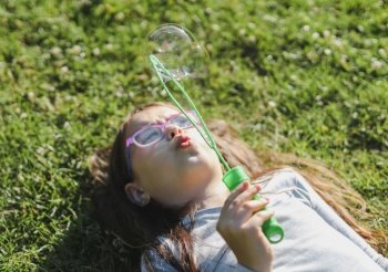Portrait of one beautiful Caucasian brunette woman with glasses, emotionally blowing soap bubbles, lying on the lawn in the park at the playground, close-up side view with selective focus. PARKS and RECREATION concept, happy childhood, children’s picnic, happy childhood, outdoor recreation, playgrounds.. Portrait of a beautiful Caucasian girl blowing soap bubbles while lying in the park.