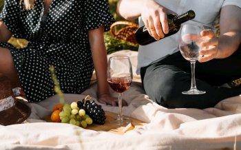 Love story, romantic picnic, valentine’s day concept. Close-up of man pouring red wine into glass while sitting with woman on blanket with fruit outdoors. Young love couple on picnic in nature.