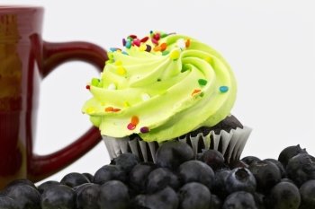 Close up of chocolate cake cupcake with green frosting in sharp focus placed with red mug in background and blueberries in foreground in shallow depth of focus