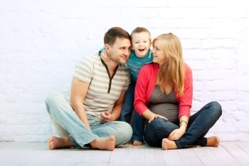 Happy family of three sitting on the floor near the wall:  mother, father and little boy. Mother is pregnant. Son is wondered.