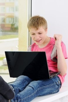 The joyful teenager with a computer sits on a window