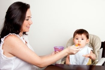Beautiful happy mother or nanny feeds funny baby boy girl orange puree with spoon, infant eats messy, while sitting at table.