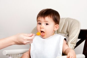 Beautiful happy baby infant boy girl with mouth open eating messy orange puree from spoon.
