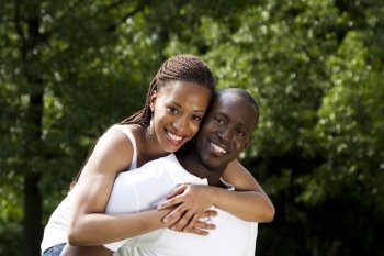 Beautiful happy smiling young African American couple in love wearing white shirts, woman hugging man, with trees in the background.