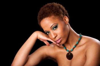Beautiful face of an African American female fashion model with bare shoulders, eyelids with green eyeshadow and long eyelashes, supporting her head with hand, and wearing a green turquoise necklace with brown wooden pendant, isolated.