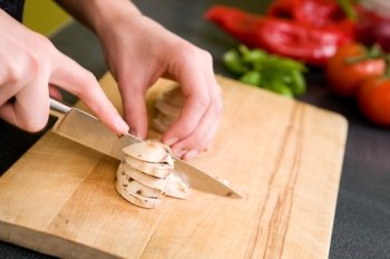 A detail image of a female hand slicing mushrooms for a pizza.  Focus is on the hand and mushrooms while tomatoes and peppers are out of focus in the background.