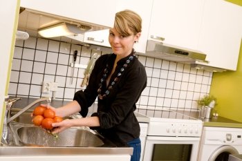 A woman is washing tomatoes in the sink at home in the kitchen.  The model is looking into the camera.
