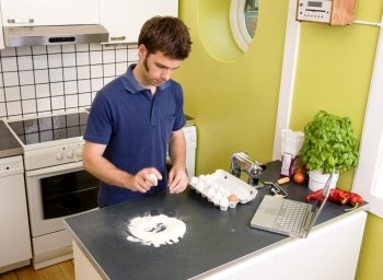 A young man making pasta at home in an apartment kitchen using a recipe from the computer