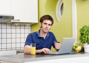 A young male shops online with a credit card in his kitchen.