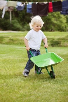 Cute young baby boy pushing a wheelbarrow in garden with clothes hanging in the background