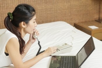 happy young asian woman working on phone and laptop in bedroom