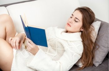 Smart woman in white sweater lying on sofa and reading book