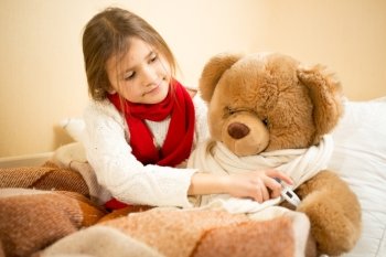 Little girl lying in bed and measuring teddy bears temperature with thermometer