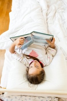 Portrait of little girl lying at bed in morning and reading big book