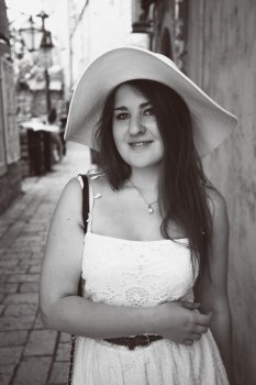 Black and white closeup photo of smiling woman wearing hat walking on narrow street