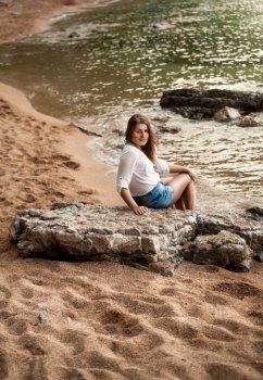 Young sexy woman sitting on big rock at sandy sea shore