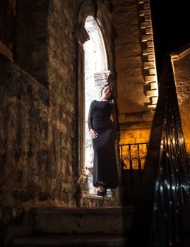 Sexy slim woman in long dress leaning against old stone arch at night