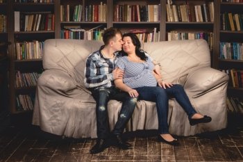 Beautiful young pregnant couple hugging on sofa against bookshelves