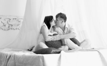 Black and white shot of happy pregnant couple kissing on bed with baldachin