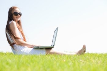 A cute young female sit on the grass in the park using a laptop