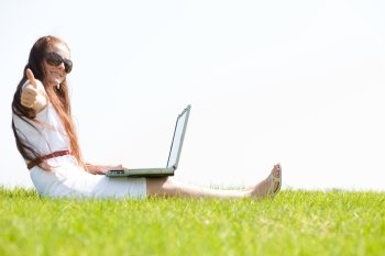 A cute young female sit on the grass in the park using a laptop