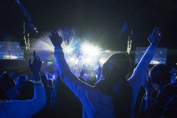 Anonymous Woman with Arms Up in Crowd of People Looking Towards Brightly LIt Stage