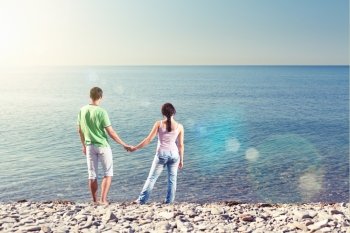 Young couple is standing near water, holding each others arms and looking over the horizon, view from behind, lens flares