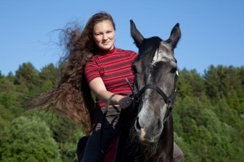 Beautiful girl with brown hair on a black horse against a blue sky and the forest