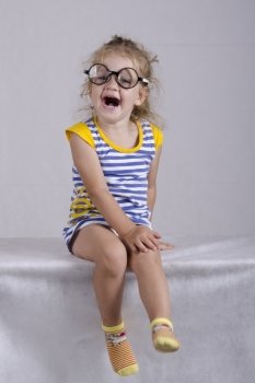 Studio, two-year-old girl sitting on the table, covered with a white cloth, and looking at the picture. Girl dressed funny glasses, laid hands on her knees and provocatively laughs