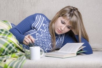 Girl sitting on the couch reading a book. Standing next to a Cup of tea, girl prevents sugar with a spoon. A girl dressed in a warm sweater and covered her warm blanket