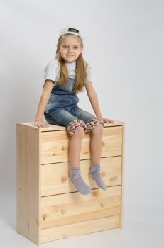 Little girl - collector of furniture in overalls relies on a chest of drawers and shows class. Girl in overalls collector relies on the chest shows class