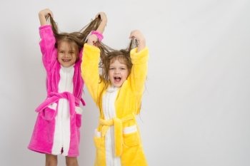 Two sister girls with wet hair standing in the bath robes on a light background. Two girls in the bath robes raised their wet hair