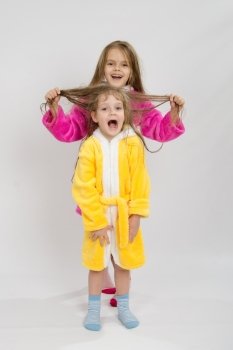 Two sister girls with wet hair standing in the bath robes on a light background. Girl holding her sisters wet hair
