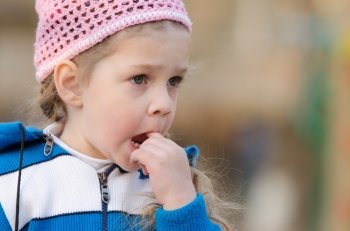 Four-year girl playing on the playground in the cool spring weather. Girl in thought stuck a finger her mouth