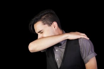 A young man sneezing into his elbow.  Black background.