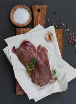 Two pieces of raw meat venison with thyme, garlic and colored pepper on a cutting board, covered with paper sheets, against the black shale stone