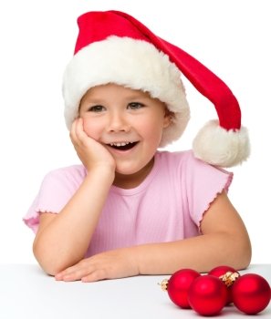 Cute little girl is sitting at table wearing santa hat, isolated over white