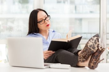 Photo of caucasian businesswoman reading a book in the office with legs on the desk