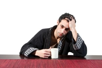 Handsome stressed guy in the morning who just woke up sitting at a table in his robe with a cup and hand in his hair, isolated on white